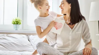 Mother and daughter sitting on a bed, holding each other with a card with a heart on it in the daughter's hands.