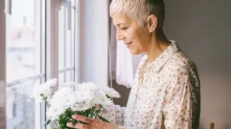An adult caucasian woman arranges a vase of flowers by the window.