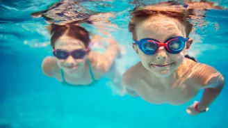 A young girl and boy swimming underwater in a pool while wearing goggles.