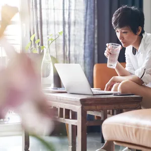 A woman drinks a cup of water while working on her laptop at home