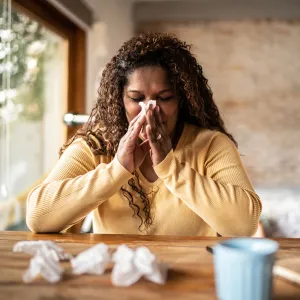 A Woman Sits at a Table with a Cup of Tea Blowing Her Nose
