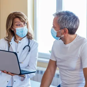 A Doctor Speaks to Her Patient in an Exam Room While Going Over His Charts on a Laptop.