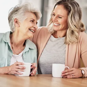 Adult woman with her arm around her mother, both smiling and holding white mugs.