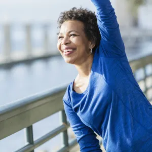 A woman is exercising outdoors, stretching her arm over her head, near the water.