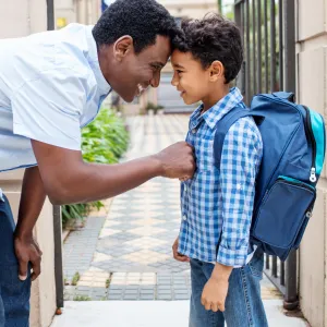 A father and a son standing outside,. The son is wearing a backpack and the father is bending over, comforting him.