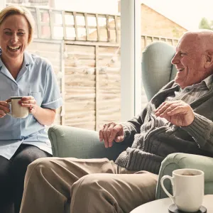 A senior man, reclined in a chair, laughs with his caretaker, who is seated next to him.