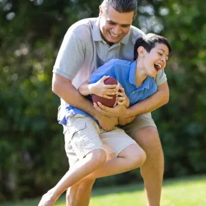 A father playfully tackles his son as they play football outside.