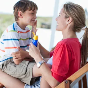 A mom with her son in her lap sitting outside in a char, applying sunscreen to his face