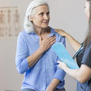 A senior woman clutching her chest, with a tech taking her blood pressure.