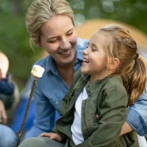 A family of four roasts marshmallows at a campground, the mother is holding her daughter as they smile.