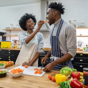 A couple snacking on veggies while cooking in the kitchen.