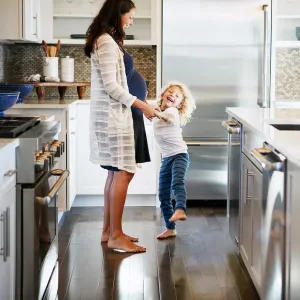 A pregnant mom and her daughter laugh in the kitchen.