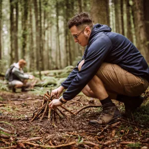 Man building a campfire in a forest while a woman collects timber in the background.