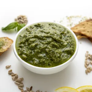 White bowl of green pesto surrounded by a piece of toast and a few crackers.