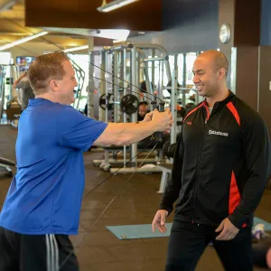 A Caucasian man exercises with a machine under the watch of his trainer.