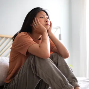 An Asian woman sits on a bed staring with a look of ennui.