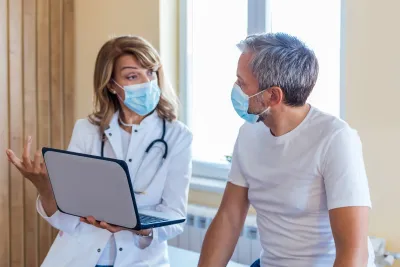 A Doctor Speaks to Her Patient in an Exam Room While Going Over His Charts on a Laptop.