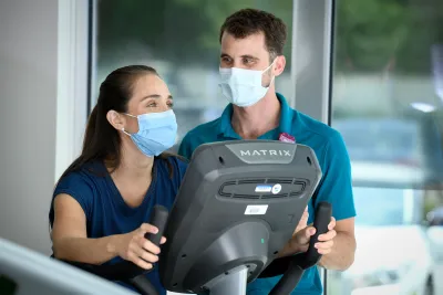 Woman riding a stationary bike while speaking with a trainer at the AdventHealth Health Park gym.