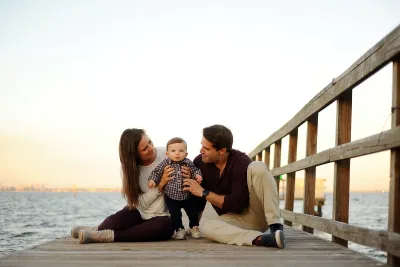 A young couple sitting on a dock holding their baby.