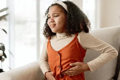 A young girl holding the left side of her abdomen in discomfort while sitting at home.