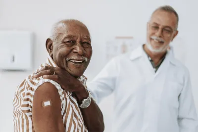 A senior man smiles and shows his arm with bandage after receiving a vaccine in the foreground while a doctor smiles in the background.