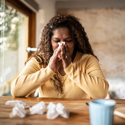 A Woman Sits at a Table with a Cup of Tea Blowing Her Nose