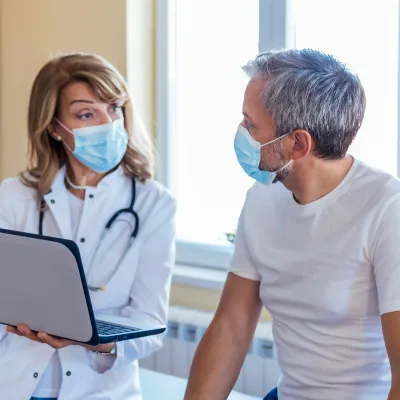 A Doctor Speaks to Her Patient in an Exam Room While Going Over His Charts on a Laptop.