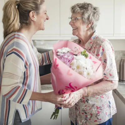 Middle aged woman giving an elderly woman light colored flowers in pink wrapping. They are both standing in a white kitchen.