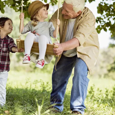 A grandfather with his two grandkids, one sitting on a wood swing, the other helping their grandfather push the swing.