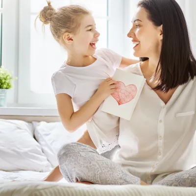 Mother and daughter sitting on a bed, holding each other with a card with a heart on it in the daughter's hands.