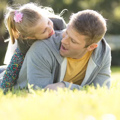 A little girl climbing on her fathers back laying down in the grass.