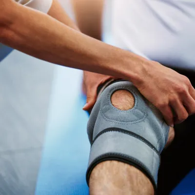 A man sitting down on a blue mat with a grey knee brace on, stretching his leg as another person helps him.