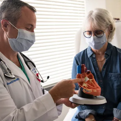 Doctor showing a woman patient a heart model while both wear masks.
