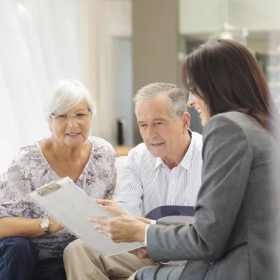 An elderly couple look hopeful as they consult with a female physician holding a clipboard.