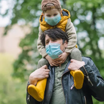 A father carrying his son on his shoulders while wearing a mask.
