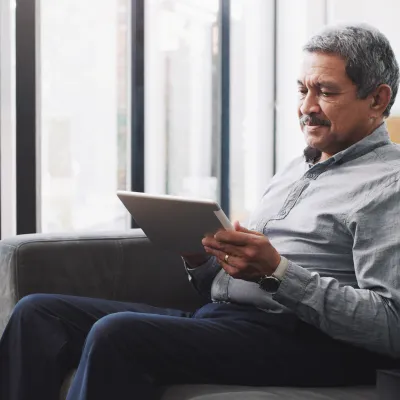 An older man sitting on a sofa reading on a tablet.