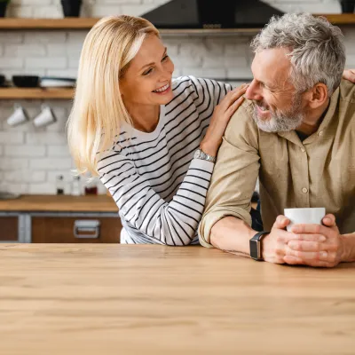 An older couple smiling at each other while in a kitchen.