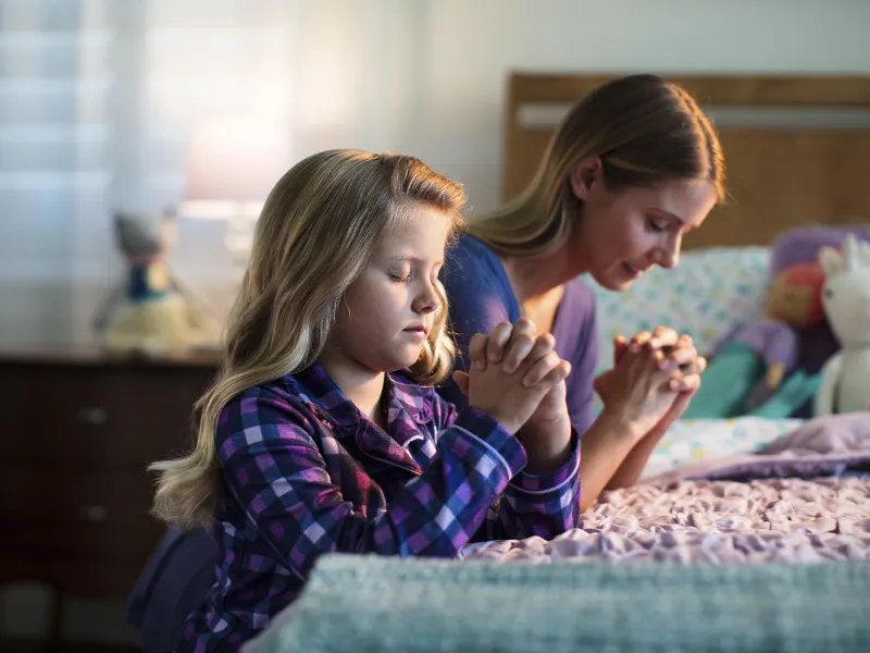 A young girl prays by her bedside with her mother before bed.
