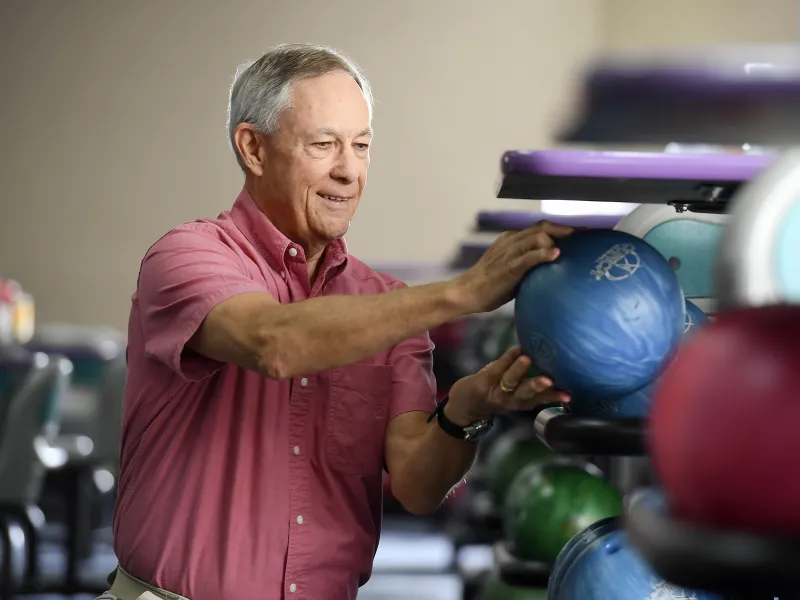 An adult man crouches to choose a bowling ball at the bowling alley.