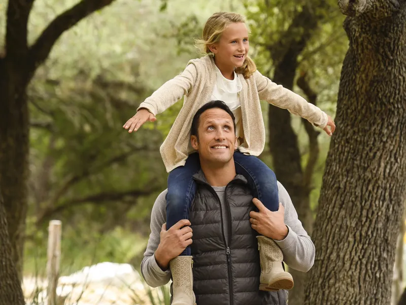 A Caucasian father carries his daughter on his shoulders while on a hike.