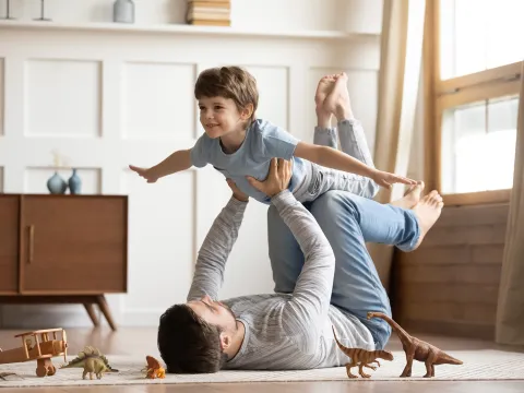 A Little Boy Plays "Air Play" on his dad's legs on the floor with dinosaur toys surrounding them