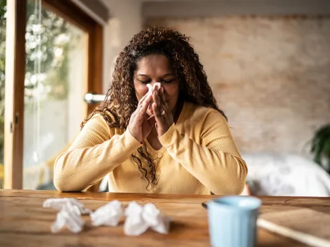 A Woman Sits at a Table with a Cup of Tea Blowing Her Nose
