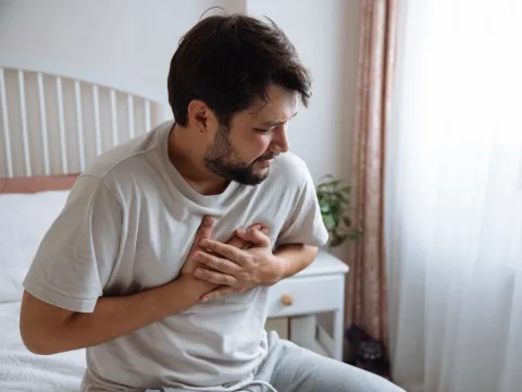 A Man Holds his Hands to his Chest While Sitting on his Bed.