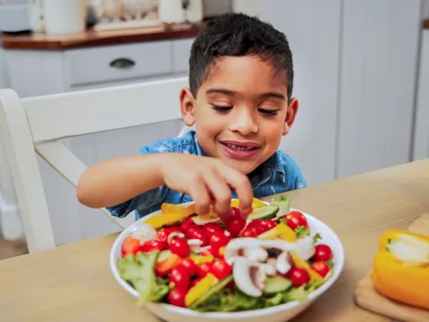 A Young Boy Grabs Some Vegetables From a Salad on a Kitchen Table.