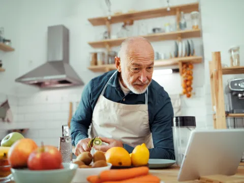 A Senior Reads A Recipe From a Tablet While Chopping Vegetables in His Kitchen