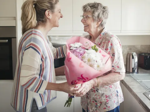 Middle aged woman giving an elderly woman light colored flowers in pink wrapping. They are both standing in a white kitchen.