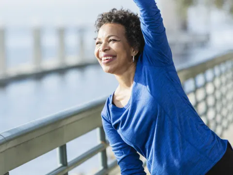 A woman is exercising outdoors, stretching her arm over her head, near the water.