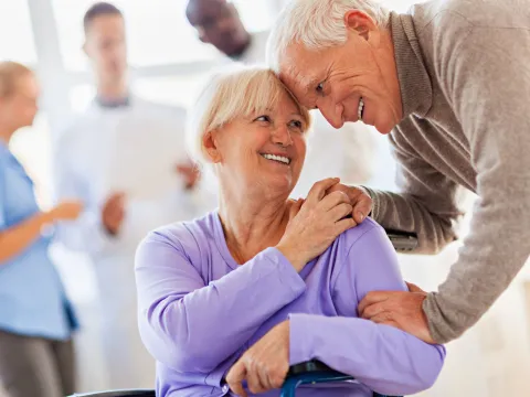 Elderly couple, the woman is sitting in a wheelchair and the man is bending down, his hands on her shoulders, comforting her.