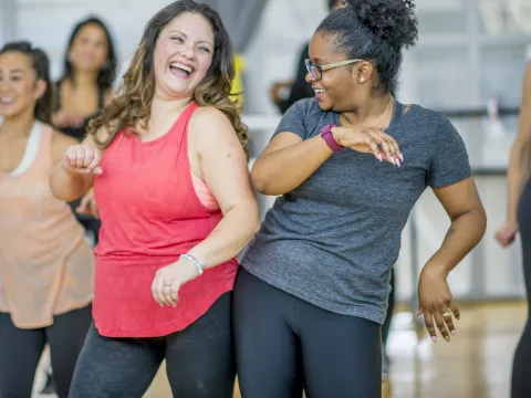 A group of adult women in an exercise class, with two women at the forefront of the shot bumping hips.