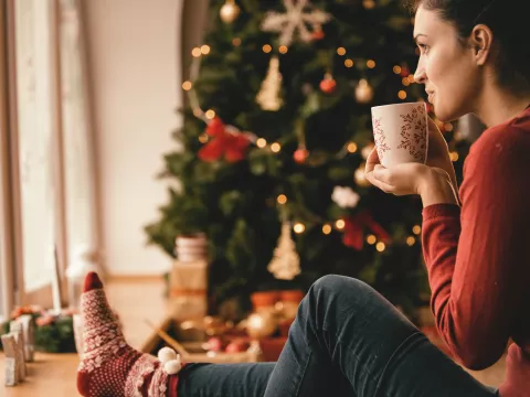 Woman sitting on the ground next to a Christmas tree, drinking something from a mug.